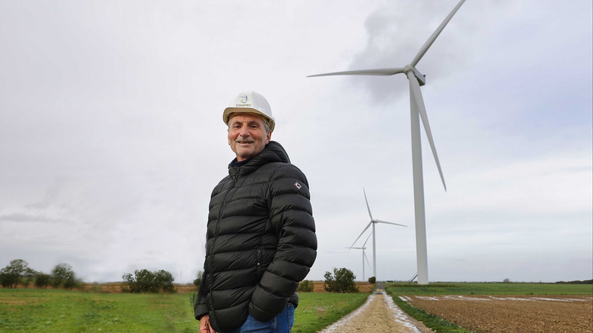 le responsable devant l'éolienne avec un casque de chantier sur la tête et en arrière plan les éoliennes